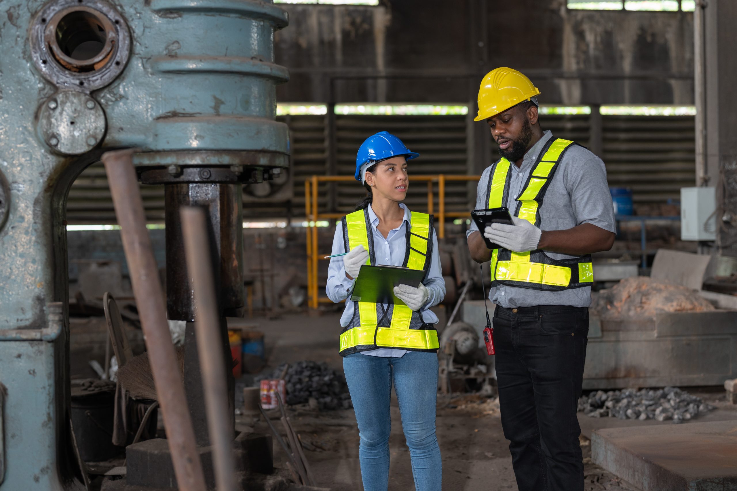 African American male and female engineers in safety vest and helmet checking and repairing old machine at heavy metal industrial factory. Preventive Maintenance concept