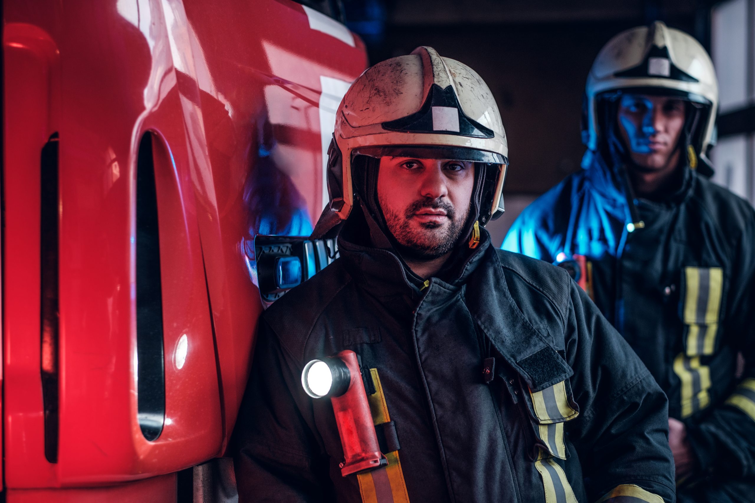 Two firemen standing near the fire truck at night in a fire depot